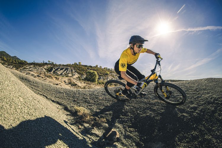 A cyclist in a yellow shirt and helmet rides a mountain bike down a gravel slope under a bright blue sky. The sun shines brightly overhead, illuminating the rugged terrain and surrounding landscape.