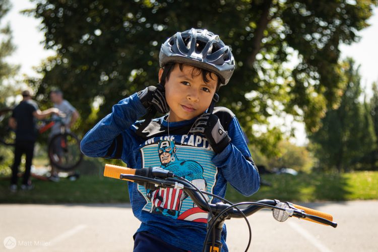 A young boy wearing a bicycle helmet and protective gloves poses confidently on his bike. He is dressed in a blue long-sleeve shirt featuring a superhero graphic. In the background, there are other people engaged with bicycles in a sunny outdoor setting with trees.