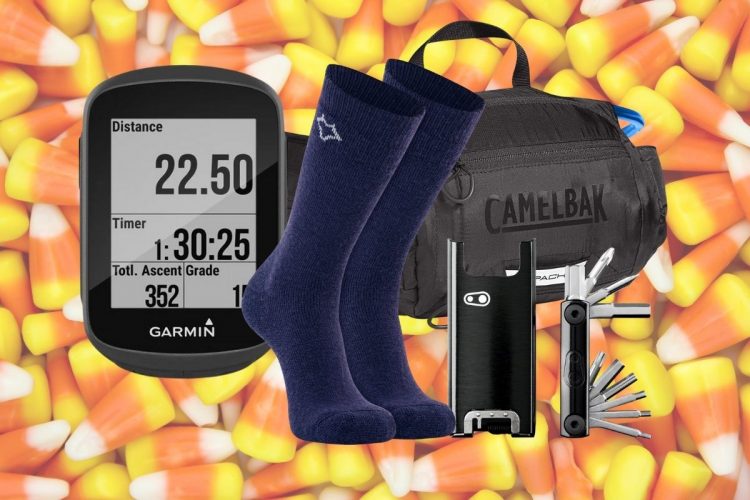 A collection of outdoor gear arranged over a colorful background of candy corn. The items include a Garmin cycling computer displaying distance and time, a pair of navy socks, a black CamelBak bag, a multi-tool, and a compact bike tire inflator.