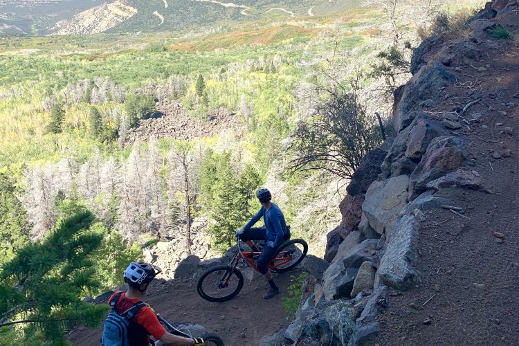 Two mountain bikers navigating a rocky trail on a hillside surrounded by trees, with a scenic valley and distant mountains visible in the background. The sky is clear, showcasing a bright and sunny day.