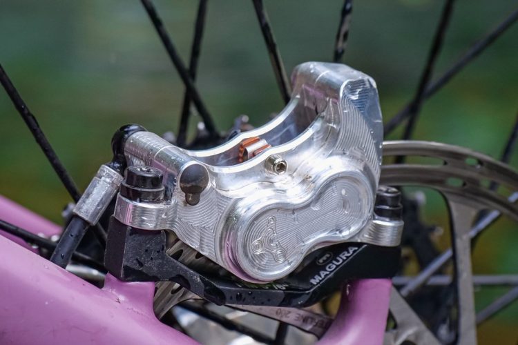 Close-up of a bike's hydraulic brake caliper mounted on a pink frame, showcasing the shiny aluminum body and intricate detailing of the components. The caliper is positioned near the rear wheel, with water droplets highlighting its design against a blurred green background.