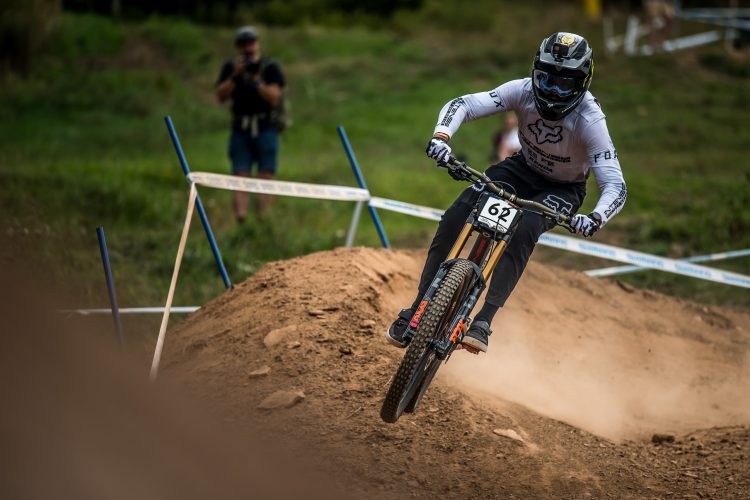 A mountain biker wearing a helmet and protective gear is mid-air, performing a jump over a dirt ramp during a competition. The biker’s jersey features the number 62, and there are spectators in the background. The scene is set in a grassy area, surrounded by race markers and a natural landscape. Dust is being kicked up from the ground as the biker lands.