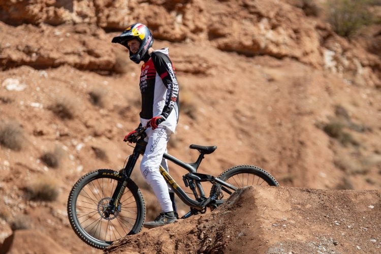 A mountain biker wearing a helmet and protective gear stands next to his bike, positioned on a dirt ramp in a rugged outdoor setting. The background features rocky terrain with sparse vegetation, highlighting an environment suitable for extreme biking.