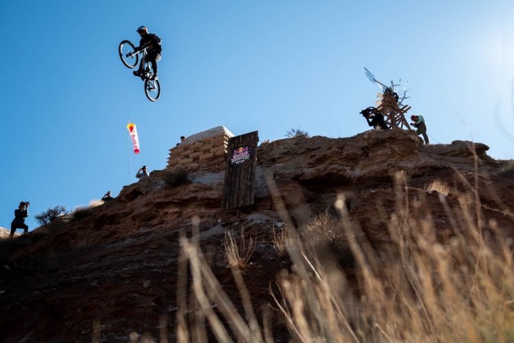 A mountain biker performing a mid-air jump from a rocky ledge, with a clear blue sky in the background. In the foreground, dry grass partially frames the image, while photographers capture the action from a nearby ridge. A Red Bull banner flutters in the breeze, indicating the event's sponsorship.