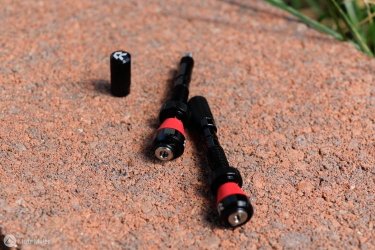 Two black and red tools with a cylindrical shape and distinct ends are positioned on a textured, reddish-brown surface. A small black cap sits nearby. The tools appear to have mechanical design features, suggesting they may be used for adjusting or fixing purposes.