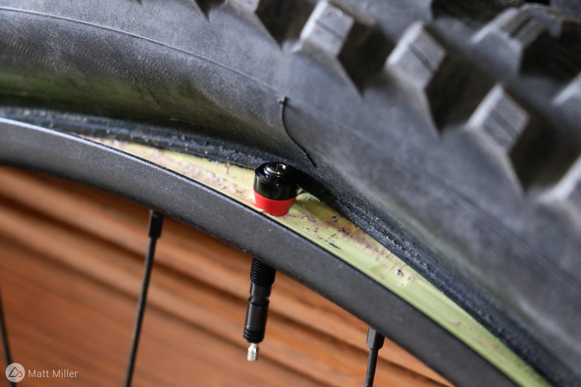 Close-up view of a bicycle tire mounted on a wheel, highlighting the valve stem and surrounding rim detail. The tire features a textured tread, and a black valve with a red accent is visible, indicating the inflation point. The background consists of a wooden surface, suggesting a workshop or storage environment.