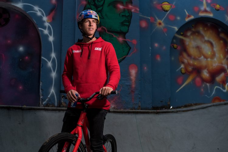 A young man wearing a red hoodie and a helmet stands on a red bicycle in a skate park. He is holding the handlebars and looking thoughtfully into the distance, with vibrant graffiti art featuring cosmic themes and abstract designs in the background. The scene is well-lit, highlighting both the rider and the colorful mural.