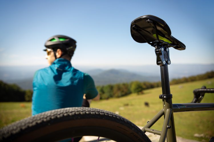 A cyclist in a blue jacket and helmet sits on a grassy hill, facing a scenic landscape of rolling hills and mountains under a clear blue sky. The foreground features a close-up view of the bike's saddle and rear wheel, emphasizing the bicycle's design.