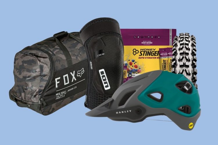 A collection of outdoor gear and accessories including a camo duffel bag from Fox, a knee protector, a teal and gray cycling helmet from Oakley, a package of Honey Stinger rapid hydration mix, and a mountain bike tire. The background is a light blue.