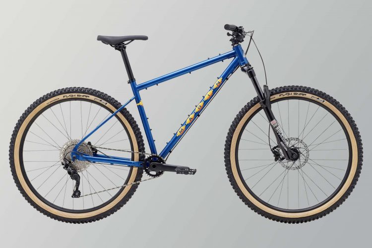A blue mountain bike with a sleek frame, featuring thick, knobby tires and a visible rear derailleur. The bike has a black front fork and a minimalist design, suitable for off-road cycling. The background is a light gradient, enhancing the bike's features.