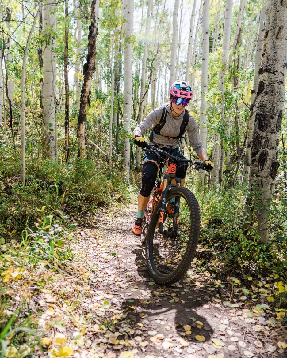 A mountain biker navigates a narrow trail surrounded by tall trees in a lush forest. The rider is wearing a helmet and protective gear, smiling as they ride through a landscape dotted with fallen leaves. Sunlight filters through the trees, creating dappled light on the path.