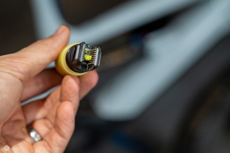A close-up image of a hand holding a small electronic component with a yellow base and multiple connecting pins. The background is blurred, showing part of a bicycle frame.