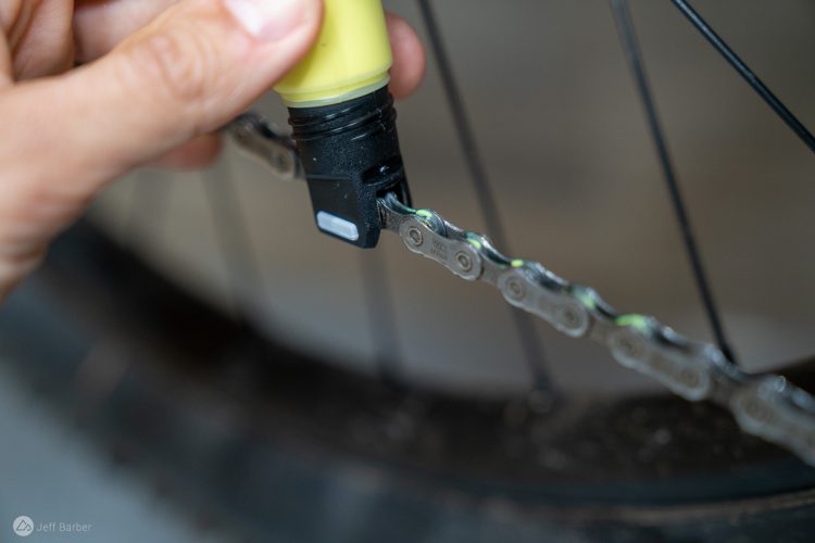 A close-up image of a person's hand applying lubricant to a bicycle chain using a yellow and black lubricant applicator. The background is blurred, focusing on the chain links, which show a slight sheen from the lubricant being applied.