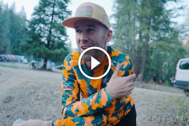 A person with a beard wearing a bright orange and teal camouflage long-sleeve shirt and a tan cap is sitting outdoors, smiling and speaking. In the background, there are trees and vehicles, indicating a nature or outdoor event. A play button overlay suggests that the image is part of a video.