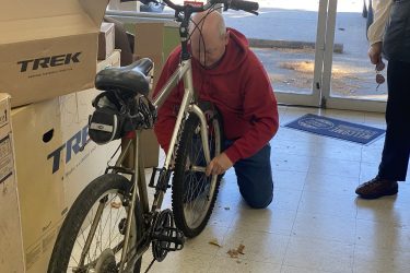 A person in a red hoodie kneels on the floor, inspecting a bicycle while using a tool. The bicycle is surrounded by cardboard boxes labeled "Trek." Another person stands in the background near the entrance of the shop. Natural light is coming through the glass doors, illuminating the scene.