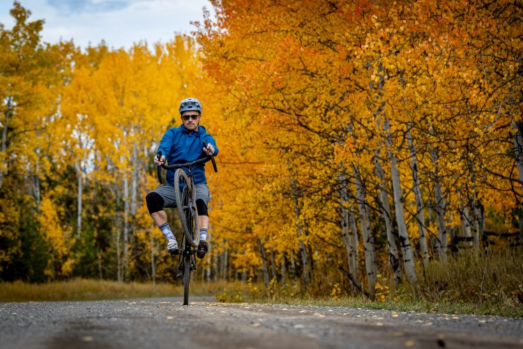 A person riding a bicycle on one wheel along a gravel path, surrounded by vibrant autumn foliage with yellow and orange leaves. The individual is wearing a helmet and a blue jacket, demonstrating a stunt as they balance on the bike.