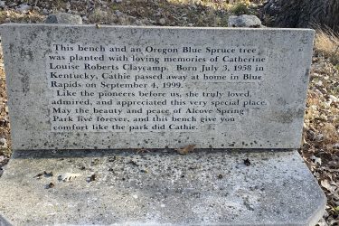 A stone bench with an engraved memorial plaque commemorating Catherine Louise Roberts Claycamp, born July 3, 1958, and passed away on September 4, 1999. The plaque expresses love for Alcove Spring Park and wishes for its beauty and peace to endure. The surrounding area features fallen leaves and natural scenery. Alcove Springs mountain bike trail.