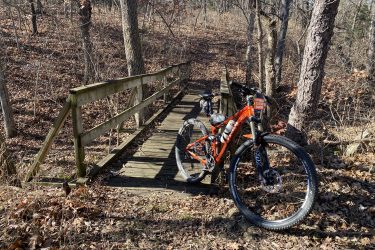 A mountain bike rests on a wooden bridge surrounded by a wooded area. The ground is covered with dry leaves, and trees with sparse foliage are visible in the background. A sign attached to the bike reads, “I’M BUILDING TO PROTECT CADE.” Drywood Creek Trail mountain bike trail.