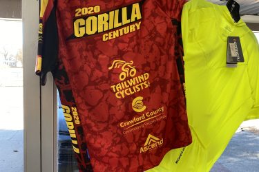 A red cycling jersey hanging on a display, featuring the text "2020 GORILLA CENTURY" prominently on the back. The jersey has yellow and black accents and logos for Tailwind Cyclists, Crawford County Convention & Visitors Bureau, and Ascend. Next to it, a bright yellow shirt is also displayed. The setting appears to be a shop with natural light coming in through a window.