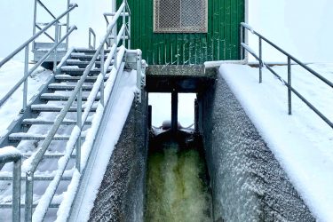 A green utility building stands atop a snowy slope, with a staircase leading up to it. Water is flowing through a channel below, creating a frothy stream. The scene is set against a light gray sky, indicating a cold, wintry day. Snow is accumulated on the ground and along the structures. Luther Marsh mountain bike trail.