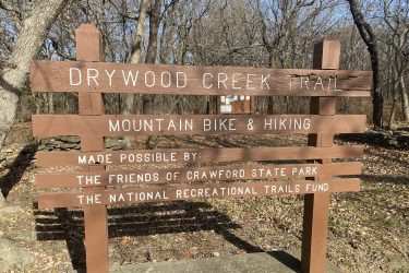 Sign for Drywood Creek Trail, indicating it is designated for mountain biking and hiking. The sign notes contributions from the Friends of Crawford State Park and the National Recreational Trails Fund. Surrounding trees indicate a natural wooded environment. Drywood Creek Trail mountain bike trail.