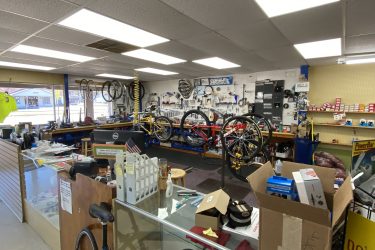Interior view of a bicycle shop featuring workbenches with tools and bike parts, neatly organized shelves with bike accessories, multiple bicycles displayed, and a counter with computers and merchandise. The shop has a well-lit atmosphere, emphasizing a focus on cycling repairs and sales.