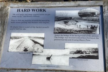 A weathered informational plaque showcasing historical techniques used for construction. The plaque features the title "HARD WORK" and a quote by Stanley Dolinar. It details the use of mule teams and slip buckets prior to 1935. Images on the plaque depict scenes of earthmoving activities, including packing down a dam, an excavator in operation, and a rainstorm affecting workers. A small inset photo shows a bridge in the background, with notes on its location relative to a marina. Drywood Creek Trail mountain bike trail.