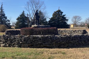 A stone monument with a statue of a figure standing atop a stone base, surrounded by a low stone wall and decorative bushes. The background features trees and a clear blue sky. The setting appears to be a park or memorial area. Drywood Creek Trail mountain bike trail.