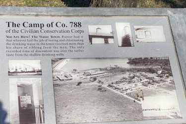 A historical plaque detailing "The Camp of Co. 788 of the Civilian Conservation Corps," featuring text about the water tower and its history, alongside several black-and-white photographs of the camp facilities and layout. Drywood Creek Trail mountain bike trail.