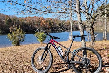 A mountain bike parked near a calm lake, surrounded by autumn foliage and bare trees. The blue sky hints at a clear day, with leaves scattered on the ground, creating a serene outdoor scene. Yonah Preserve mountain bike trail.