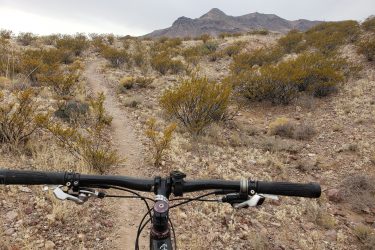 View from the handlebars of a mountain bike looking down a rocky trail, surrounded by sparse vegetation and hills under a cloudy sky. Socorro Singletrack mountain bike trail.