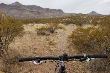 A view from the handlebars of a mountain bike on a rocky trail surrounded by dry grass and bushes, with rolling hills and mountains in the background under a cloudy sky. Socorro Singletrack mountain bike trail.