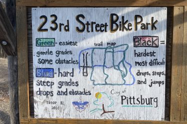 Sign at 23rd Street Bike Park showing a trail map and difficulty levels. It categorizes trails by color: Green for easiest with gentle grades and some obstacles, Blue for hard with steep grades, drops, and obstacles, and Black for the hardest, most difficult trails featuring drops, steps, and jumps. The sign is located in Pittsburg and includes troop details. 23rd St Bike Park mountain bike trail.