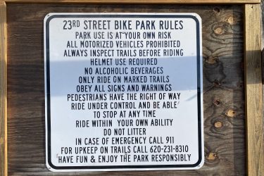 Sign outlining the rules for 23rd Street Bike Park, including safety guidelines such as helmet use, prohibition of motorized vehicles, alcohol restrictions, and the need to obey all signs. The sign emphasizes riding under control, yielding to pedestrians, and proper park etiquette. Emergency contact information is also provided. 23rd St Bike Park mountain bike trail.