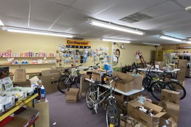 A cluttered bicycle shop interior filled with various bike parts and accessories. Several bicycles are displayed among stacks of cardboard boxes and products, with a wall showcasing organized items such as tires and tools. The atmosphere appears busy and in the process of restocking.