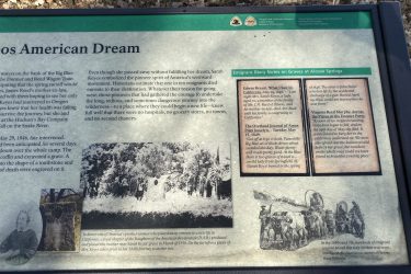 Informational plaque about the experiences of pioneer women on the Oregon Trail, detailing the journey of Sarah Keyes and her challenges, including her death before reaching her destination. The plaque includes excerpts from diaries of other pioneers, historical context, and a tribute to Keyes, with a photograph of her grave marker. The setting is outdoors, surrounded by trees and grass. Alcove Springs mountain bike trail.