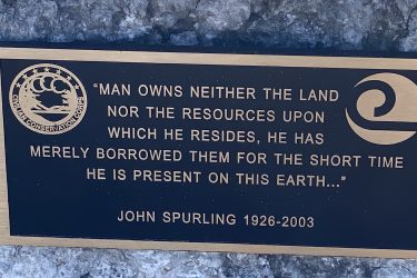 A bronze plaque featuring a quote about the relationship between man and the land, attributed to John Spurling (1926-2003). The inscription reads: "Man owns neither the land nor the resources upon which he resides, he has merely borrowed them for the short time he is present on this earth..." The plaque also displays the emblem of the Civilian Conservation Corps. The plaque is mounted on a rocky surface. Drywood Creek Trail mountain bike trail.