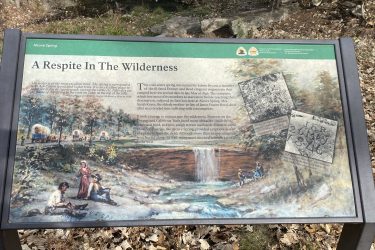 A sign titled "A Respite In The Wilderness" at Alcove Spring, featuring historical information about the spring's significance to pioneers along the Oregon and California Trails. The sign includes a painted scene depicting travelers resting by a water source, surrounded by trees and a waterfall. Historical notes discuss the hardships faced by emigrants and mention specific individuals connected to the site. The background shows rocks and a natural landscape. Alcove Springs mountain bike trail.
