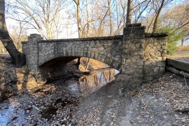 A stone arch bridge spanning a shallow creek, surrounded by barren trees and a landscape covered with fallen leaves. The scene is set on a sunny day with clear blue skies. Cottonwood River Trail mountain bike trail.