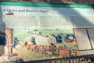 A historical interpretive sign titled "Alcove Spring," describing the challenges faced by emigrant wagon trains in the mid-1800s as they journeyed to Oregon and California. The sign features text detailing the experiences of pioneers, including heavy spring thunderstorms and crowded conditions at crossings. It includes a colorful illustration of a serene landscape with wagons, oxen, and figures engaging in daily chores. The background highlights the natural beauty and importance of Alcove Spring as a rest stop during westward expansion. Alcove Springs mountain bike trail.