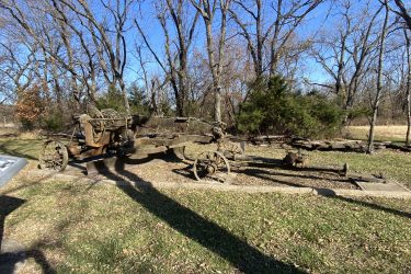 An old, rusted agricultural implement is positioned on a stone base surrounded by bare trees and grass. The implement has large wooden wheels and appears to be in a state of disrepair, highlighting its historical significance. In the foreground, a metal sign is partially visible, providing context about the artifact. The scene is set against a clear blue sky, indicating a sunny day. Drywood Creek Trail mountain bike trail.