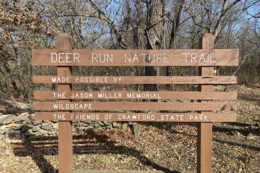 A wooden sign marking the entrance to the Deer Run Nature Trail, with text indicating it was made possible by the Jason Miller Memorial, Wildscape, and the Friends of Crawford State Park. The sign is surrounded by a natural setting, with trees and fallen leaves visible in the background. Drywood Creek Trail mountain bike trail.