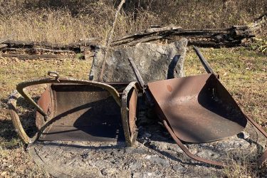 Two old, rusted wheelbarrows are positioned on a circular stone base surrounded by dry grass and sparse foliage. In the background, a weathered wooden fence can be seen, adding to the rustic setting. The scene conveys a sense of abandonment and decay in a natural environment. Drywood Creek Trail mountain bike trail.