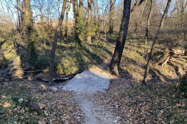 A narrow dirt path leading through a wooded area with bare trees and sparse underbrush. A small concrete slab is visible on the path, with scattered fallen leaves surrounding it. The sunlight highlights the trees and the terrain, creating a serene nature scene. Cottonwood River Trail mountain bike trail.