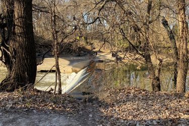 A serene view of a river flowing past a small concrete dam, surrounded by bare trees and autumn leaves on the ground. The setting captures the tranquility of nature in late fall, with sunlight filtering through the branches. Cottonwood River Trail mountain bike trail.