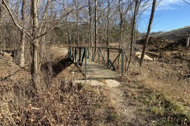 A small wooden bridge with a green metal railing spans a narrow ditch in a wooded area. Bare trees surround the bridge, and the ground is covered with dried grass and leaves. In the background, there is a hill and some construction debris visible. The sky is clear with a few clouds. Drywood Creek Trail mountain bike trail.