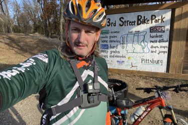 A person wearing a cycling helmet and a green jersey stands in front of a bike park sign that details the trail map and difficulty levels for different biking paths. An orange mountain bike is parked beside them. The background features wooded terrain. 23rd St Bike Park mountain bike trail.