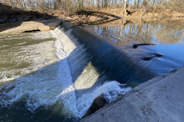 Water cascading over a concrete dam into a river, surrounded by bare trees and a clear blue sky. The water creates ripples and splashes as it flows downstream. Cottonwood River Trail mountain bike trail.