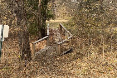Wooden bridge crossing over a path covered in fallen leaves, surrounded by trees in a natural setting, with a directional sign indicating a creek nearby. Alcove Springs mountain bike trail.