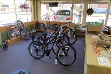 A bicycle shop interior featuring several bicycles of various styles, including mountain and children's bikes. The shop has a wooden display area and large windows letting in natural light. There are also cardboard boxes and a green armchair visible in the space.